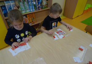 Dwoje dzieci siedzi przy stoliku i składają obrazek z części przedstawiający godło Polski.